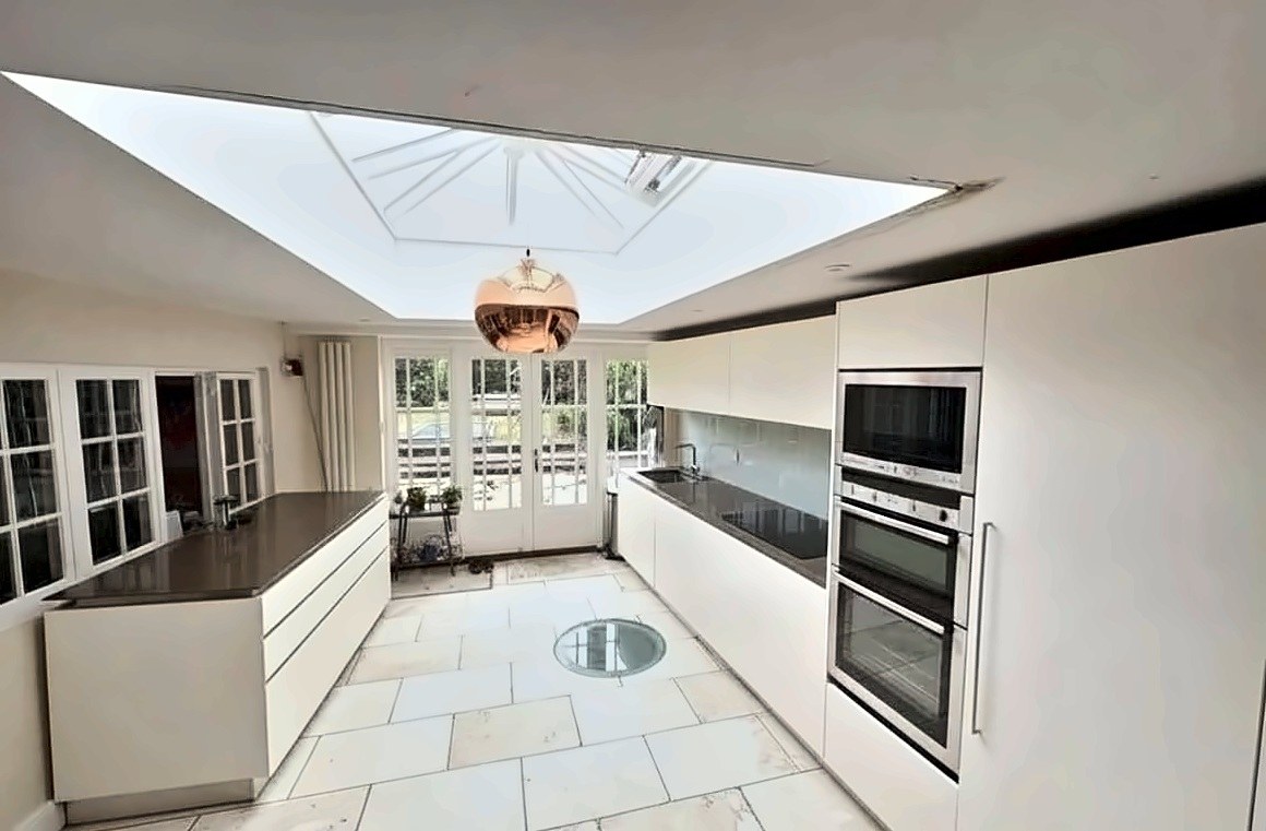 Handleless cream kitchen in an orangery extension with a lantern rooflight, copper pendant and travertine floor.